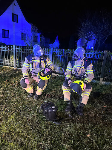 Zwei Feuerwehrkräfte mit Atemschutzgeräten sitzen nachts auf Stühlen vor einem Grundstück.