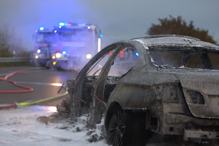 Ein ausgebranntes Fahrzeug mit Resten von Löschschaum und Feuerwehrfahrzeugen im Hintergrund.