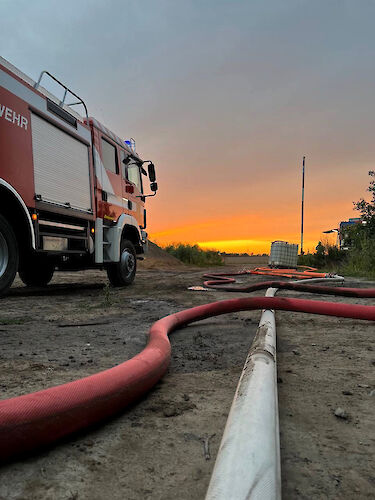 Ein Feuerwehrfahrzeug und Schläuche auf einem Sandweg vor einem Sonnenuntergang.