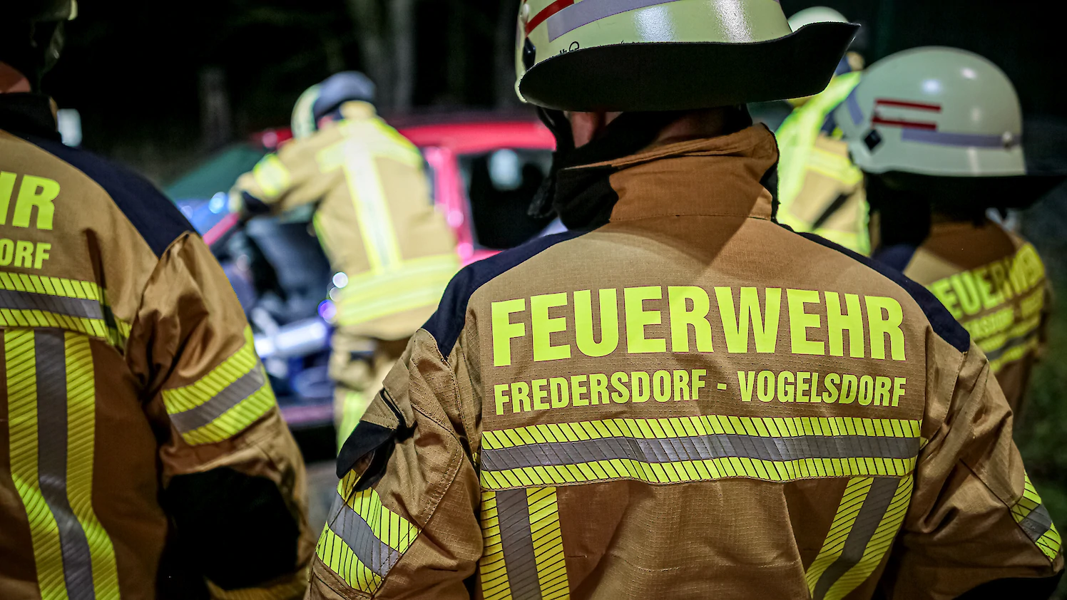 Drei Feuerwehrkräfte stehen im Halbkreis um einen Kameraden, der mit technischem Gerät ein Auto öffnet.