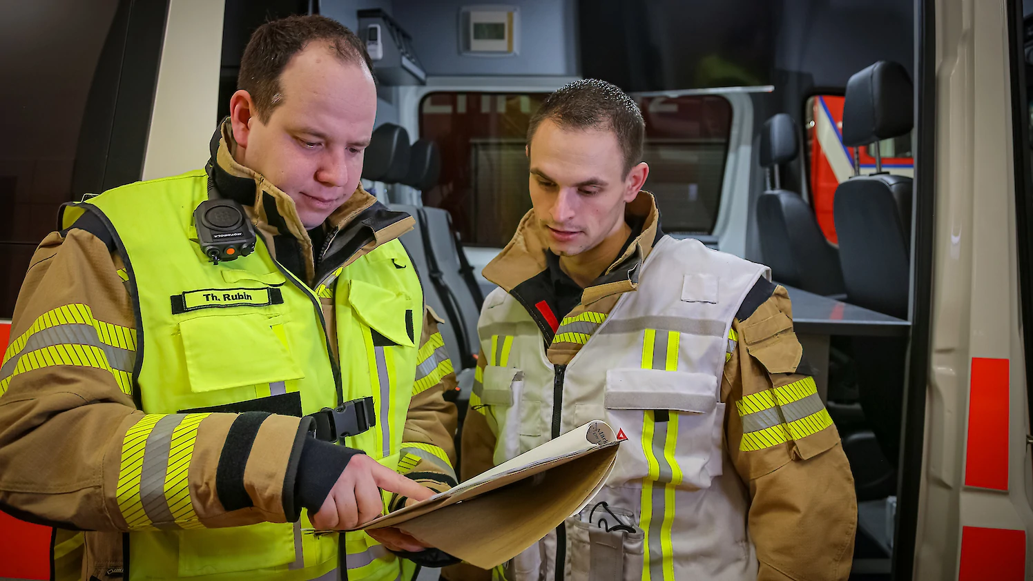 Zwei Feuerwehrmänner mit Funktionswesten lesen in einer Arbeitsmappe.