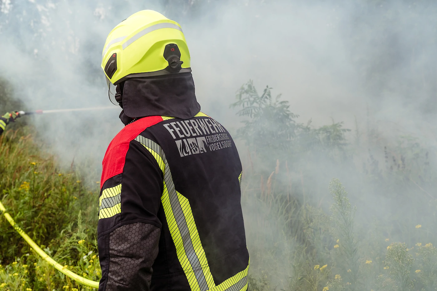 Eine Feuerwehrkraft vor einer verqualmten Wiese, gerade von einer anderen Person gelöscht wird.