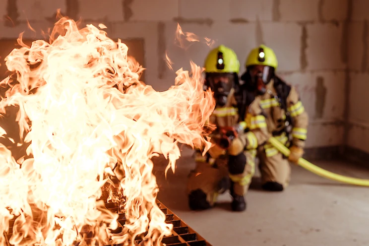 Zwei Feuerwehrmänner mit Atemschutzgeräten und einem Schlauch knien vor einem Feuer in einem Gebäude.